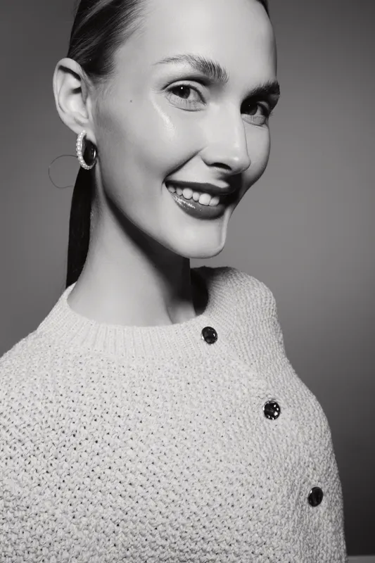 Smiling white woman with light skin and long hair wears knitted sweater and hoop earrings, posing against grey background. Concept of beauty and fashion portrait.