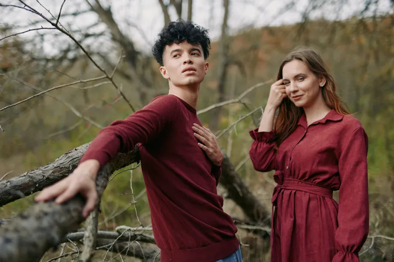 A candid pair stands in a quiet woodland, burgundy garments and relaxed pose conveying simple style and emotional depth, authenticity evident in natural expression and posture.