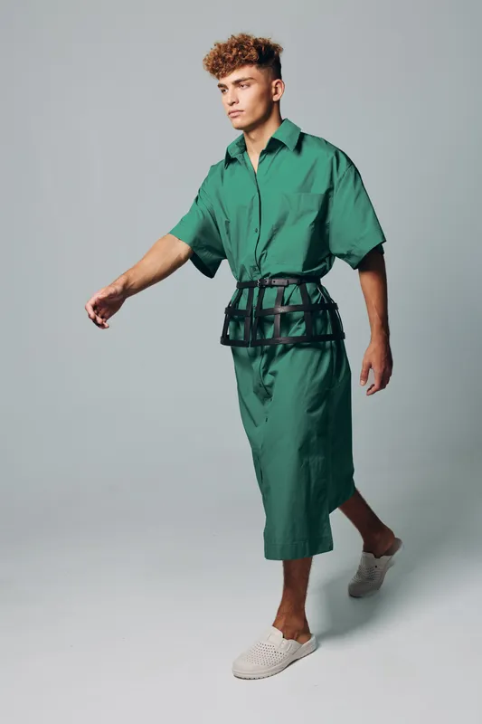 Young man walking in green oversized shirt and matching wide pants with black belt bag and white slip on shoes on gray background in studio. Casual fashion style and confident pose.