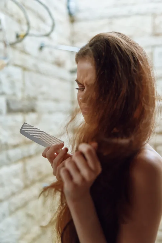 woman with tangled hair holding a comb looks frustrated in neutral bathroom setting, curly brown hair, natural light, personal care concept