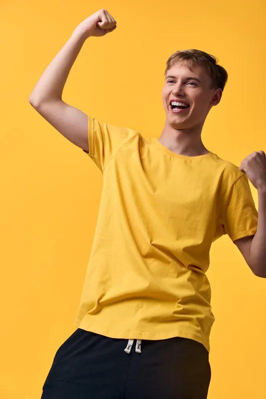 Cheerful man in a bright yellow shirt raises his fists in a victory gesture against a matching yellow backdrop, radiating energy, joy and success in a dynamic lifestyle moment.