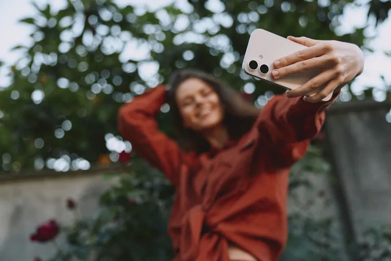 Smiling woman in a warm brown coat taking a selfie outdoors, greenery in the background, lifestyle moment, casual portrait, natural light, cheerful mood, candid photography