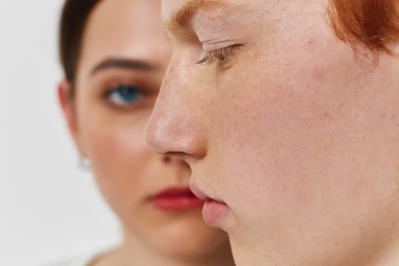 Closeup profile of young redhead man with freckles and serious expression with woman face blurred in background. Intense mood with focus on skin texture and natural features.