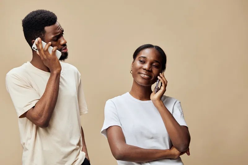 A joyful African couple chatting on mobile phones, showcasing positive emotions and modern attire against a neutral beige background