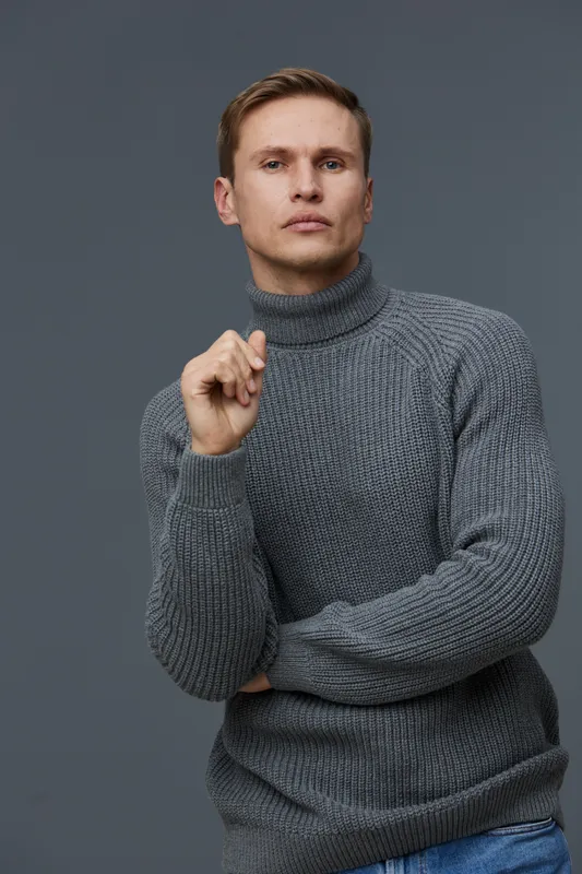 Frontal portrait of thoughtful young man with short brown hair wearing gray turtleneck sweater gazing serious at camera on plain gray background studio shot. People lifestyle concept