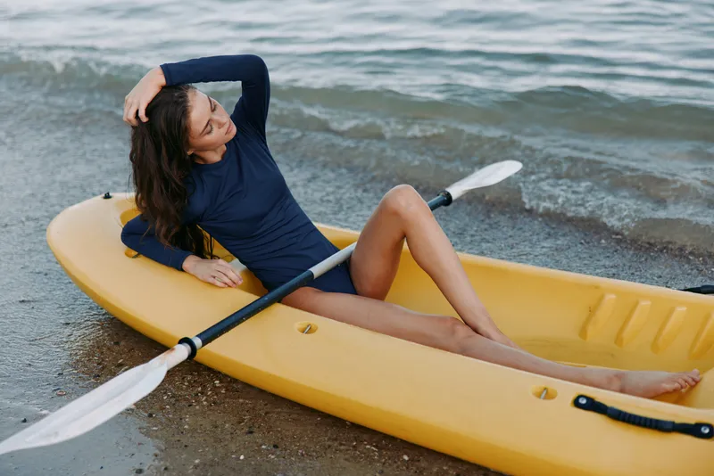 Female paddler lounges on a yellow kayak by the shoreline, arm raised, resting in a relaxed pose as gentle waves wash the sand, capturing outdoor leisure.