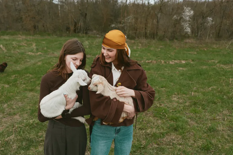 Two women in casual jackets cradle a goat kid in an open field, showcasing care, bonding, and friendship in a sunny outdoor farm setting, a candid lifestyle moment with gentle animal interaction.