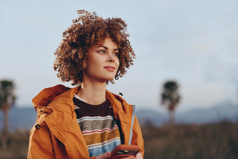 Woman lifestyle outdoors portrait, curly hair, orange jacket, calm gaze and wind, open field with distant mountains, soft sky, natural daylight