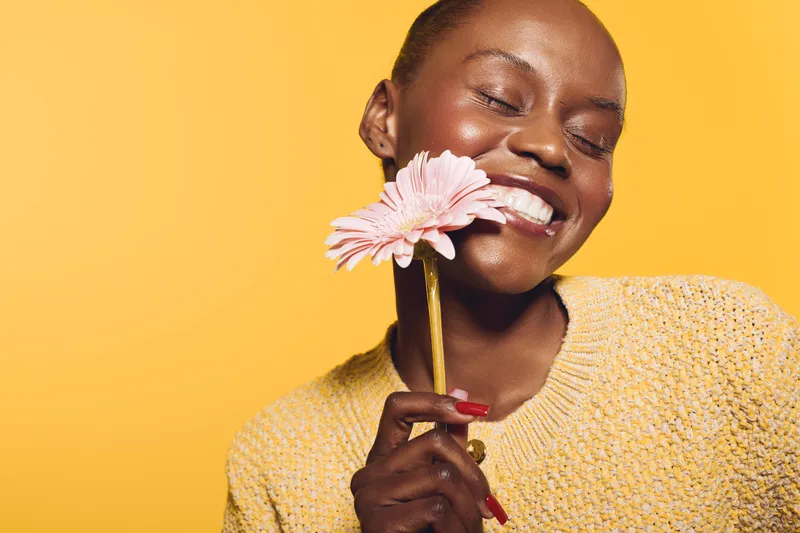 Close-up of happy African woman with dark skin holding pink flower near face, smiling with closed eyes. Bright yellow background and cozy sweater create cheerful mood and natural beauty concept.