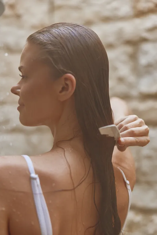 Woman combing wet hair in shower with natural stone background, relaxed and focused, displaying self-care and morning routine in soft warm lighting.