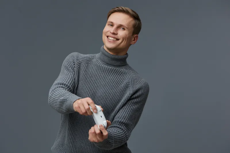 Cheerful smiling young man with short hair wearing cozy gray sweater, holding soap dispenser in hand, looks at camera, studio portrait isolated on plain gray background. People lifestyle concept, wint