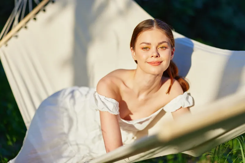 Young woman in a white dress relaxing in a hammock, smiling joyfully, with green nature background, embodying tranquility and summer vibes