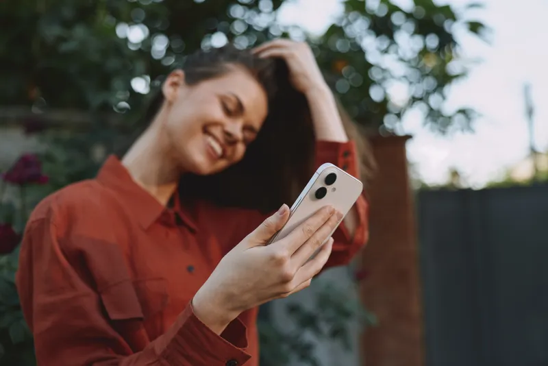 Cheerful woman outdoors, smiling while taking a selfie in a sunlit garden, casual red shirt, relaxed pose, natural colors, fresh greenery background