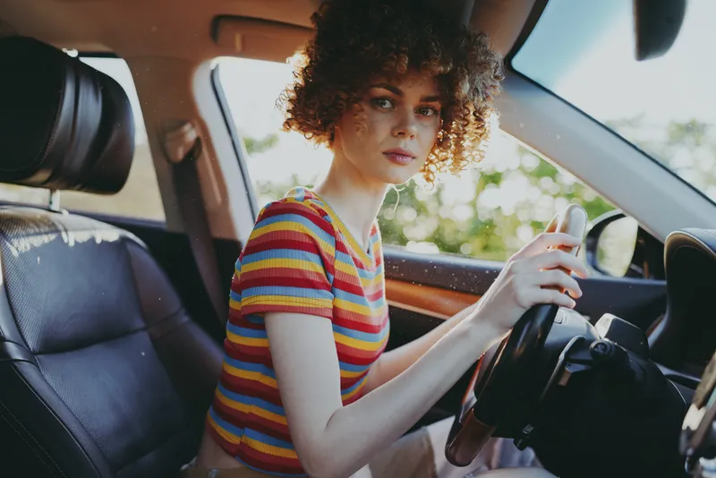 A stylish woman driver sits in a retro car with a striped top, looking at the camera. Warm film colors evoke casual lifestyle and vintage automotive aesthetics.