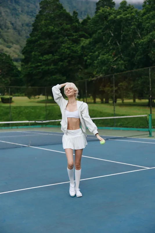 Young female athlete in a sporty white outfit joyfully playing tennis on a sunny court surrounded by lush greenery, showcasing an energetic and healthy lifestyle