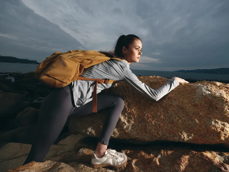 Athletic woman climbing a rock during outdoor adventure in nature, wearing sports attire and looking determined, with a backpack and dramatic cloudy sky
