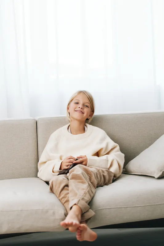 Young girl sits barefoot on a light sofa, wearing a soft cream sweater and a relaxed smile, creating a warm, homey portrait of comfort and casual charm.