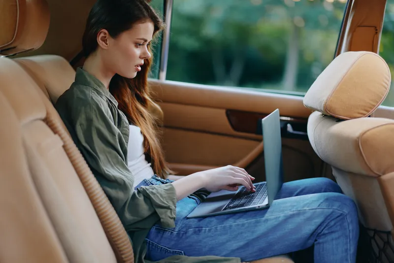 Young woman working on a laptop in the back seat of a car, dressed casually, reflecting concentration and productivity in a natural outdoor setting.