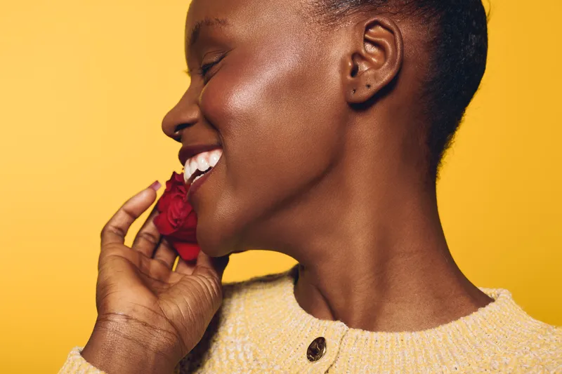 Smiling dark skinned African woman with short hair holds red rose near face, eyes closed, enjoying scent. Yellow background and soft lighting create warm, joyful mood of natural beauty and happiness.