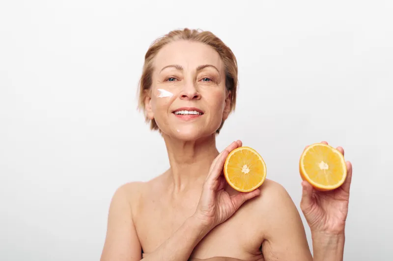 Smiling mature woman holds two halves of fresh orange fruit near face with facial cream on cheek. Concept for natural skincare, healthy lifestyle, and vitamin C beauty treatment.