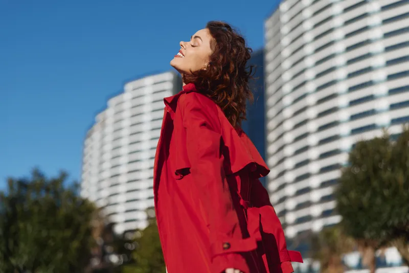 Cheerful woman outdoors in a red coat, eyes closed, basking in sunlight beside modern buildings and a clear blue sky, relaxed pose.