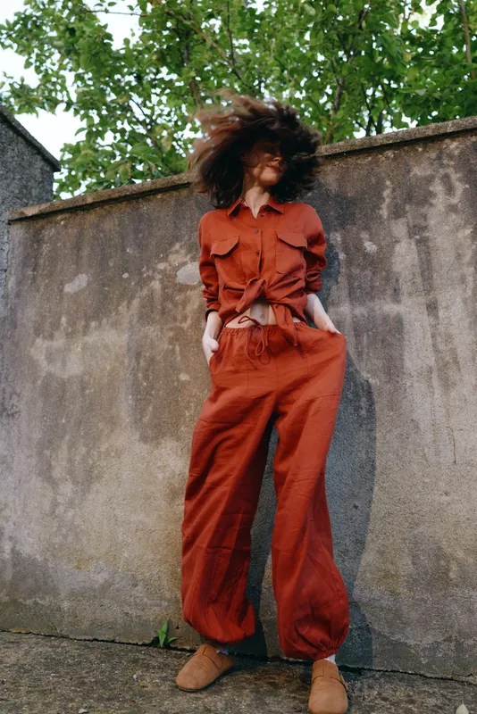 Young woman in a burnt orange jumpsuit stands against a weathered wall, wind blows her curly hair, and she poses with hands in pockets, radiating confidence