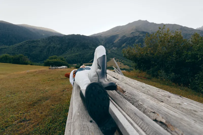 Person on bench in mountains nature relax boots meadow outdoor lying on wooden plank with jacket, peaceful landscape, distant peaks and green valley for weekend escape and rest.