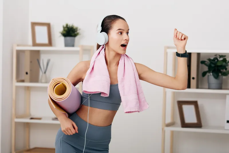 Fitness woman in a workout studio wearing headphones, carrying a yoga mat and towel, celebrating a victory pose after a workout in a home gym, energetic and motivated for sport.