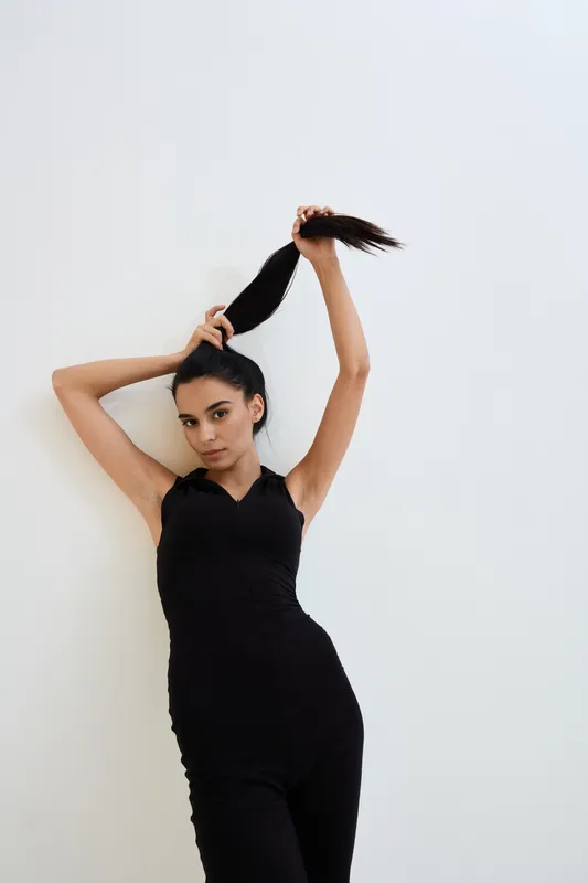 Young woman in black jumpsuit posing against white wall, holding her long dark ponytail with one hand, stylish confident expression, studio portrait with minimalistic background.