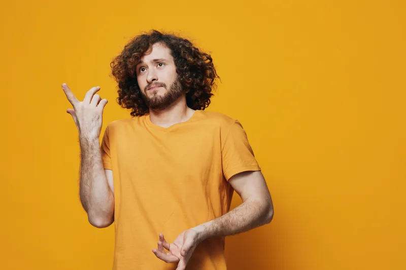 Young man expressing emotions with curly hair, wearing a casual orange t shirt, against a vibrant yellow background, showcasing joy and positivity
