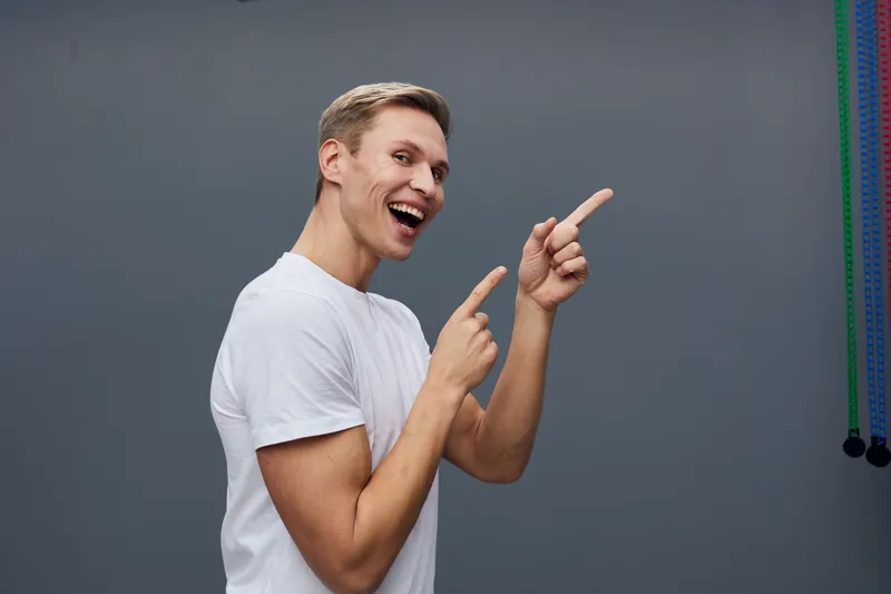 Smiling young caucasian man casual white t-shirt pointing fingers isolated dark gray background digital nomad concept lifestyle