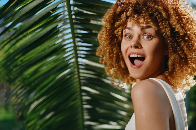 Smiling woman with curly hair posing happily outdoors near tropical green palm leaves under natural sunlight, showcasing joyful expression and vibrant summer vibe.