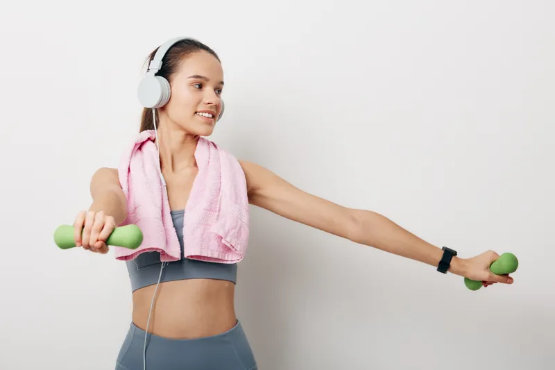 Woman fitness training with dumbbells, wearing headphones and a pink towel after workout, arms extended, gym setting, sporty apparel and motivation for exercise, strength building and healthy