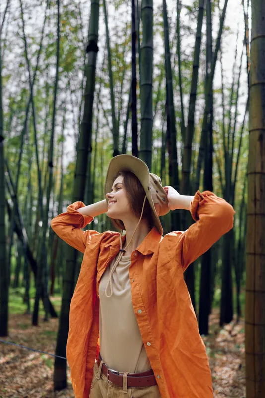 Woman in a bamboo forest wearing a hat and orange jacket, portrait shot capturing a peaceful smile, enjoying nature and outdoor travel among tall green bamboo trunks.