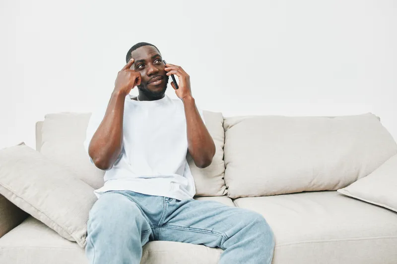 A Black man sitting comfortably on a sofa, engaged in a phone conversation, displaying curiosity and confidence, with a minimalistic white background