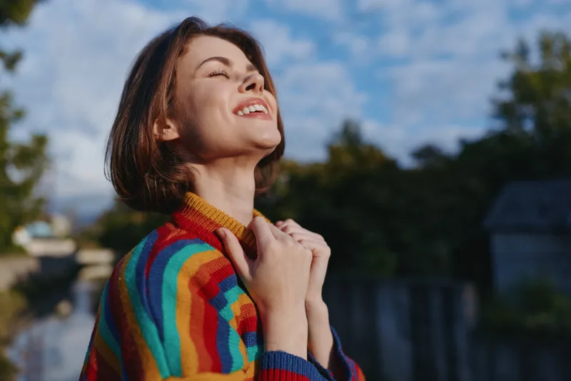 Young woman, smiling woman in rainbow sweater with eyes closed outdoors in nature by a canal, short brown hair and clasped hands, joyful expression and cozy casual outfit for travel lifestyle