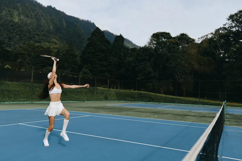 Athletic woman serving on a tennis court in a white sporty outfit surrounded by lush green mountains, showcasing determination and focus, ideal for sports and wellness themes