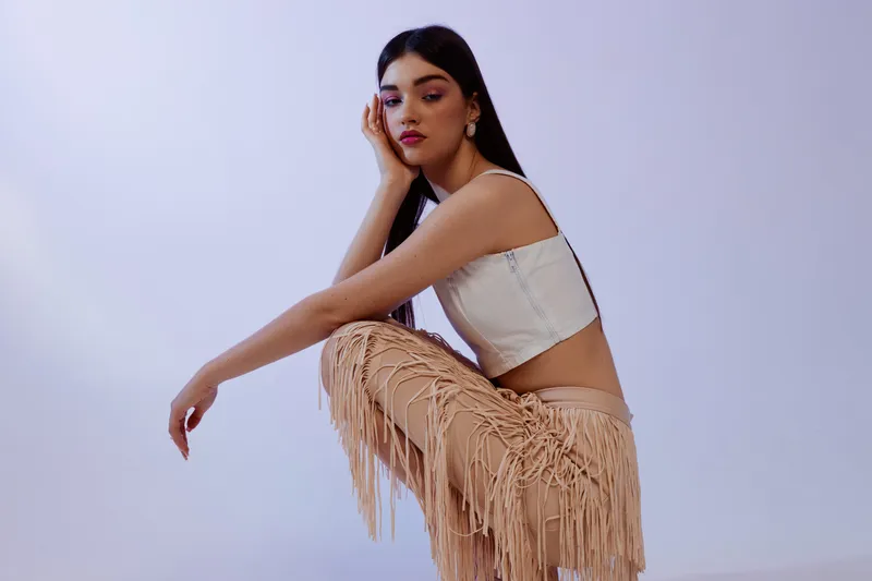 Young woman beauty fashion model poses sitting with hand on face wearing a white top and fringed pants. Studio shot on a light background. Concept for style, elegance, and confidence.