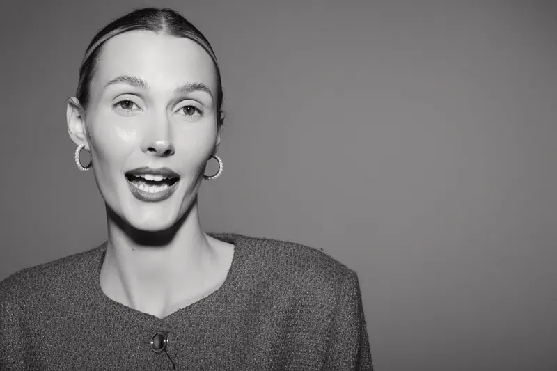 Young Caucasian woman with fair skin and slicked back hair speaks with expressive face on gray background.