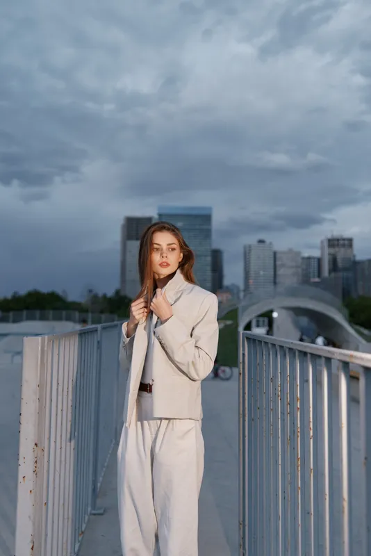 Woman wearing light beige suit standing on urban bridge with overcast sky and modern cityscape background in business fashion style