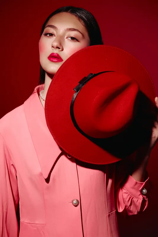 Female fashion model wearing pink blazer and red hat looks confidently at camera with bold red lipstick. Studio portrait with crisp lighting on red background symbolizes style and beauty creativity.
