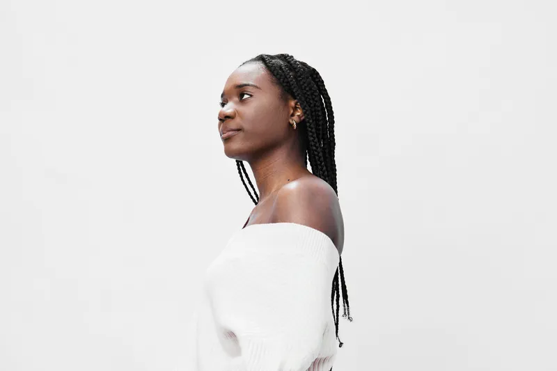 A thoughtful young woman with braids wearing a white off shoulder sweater, looking to the side against a light background Perfect for lifestyle themes