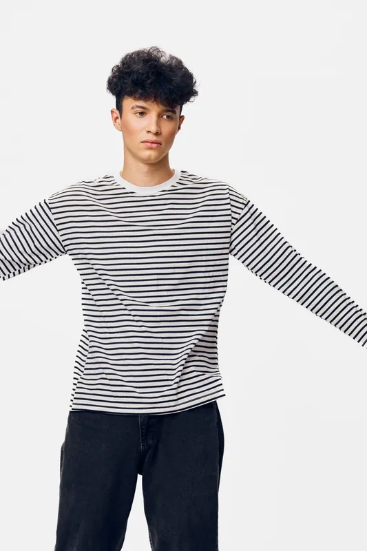 Young man with curly dark hair wearing a striped long-sleeve shirt and black pants stands with arms outstretched against a plain white background, casual and relaxed.