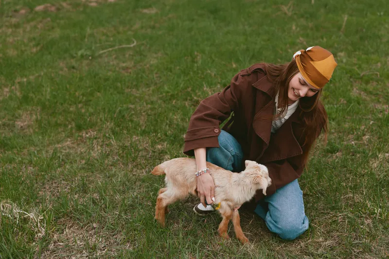 Caring woman kneels on grass, guiding a young goat kid with gentle attention. Outdoor lifestyle moment showcasing farm life, companionship, and calm connection in natural light.