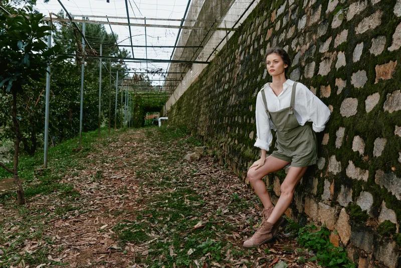 Young woman in rustic countryside setting wearing olive green overalls, white blouse, and boots, expressing a relaxed emotion surrounded by nature s greenery and earthy tones