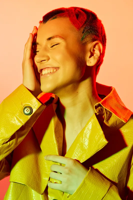 smiling woman with short hair wearing yellow coat laughs joyfully with eyes closed against warm orange background, radiating happiness and vibrant energy