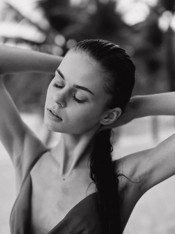 Monochrome portrait of a serene young woman with eyes closed and arms raised, hair tied back, capturing quiet grace and a moment of stillness against a soft background.