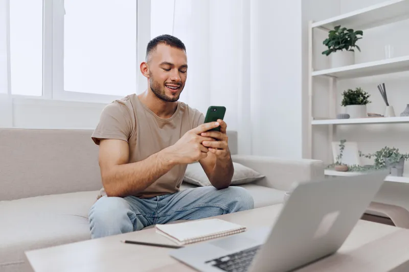 Happy man sitting on the couch using smartphone, casual attire, bright living room, modern decor, relaxed atmosphere, digital connectivity.