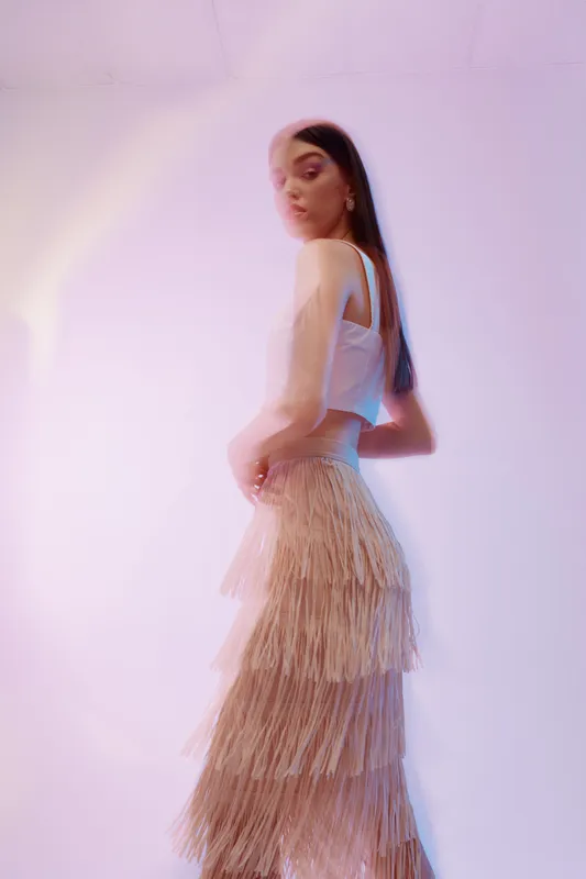 Young woman in beige fringed skirt and white top poses in soft pastel lighting. Fashion and beauty concept with elegant style and delicate mood in studio environment.