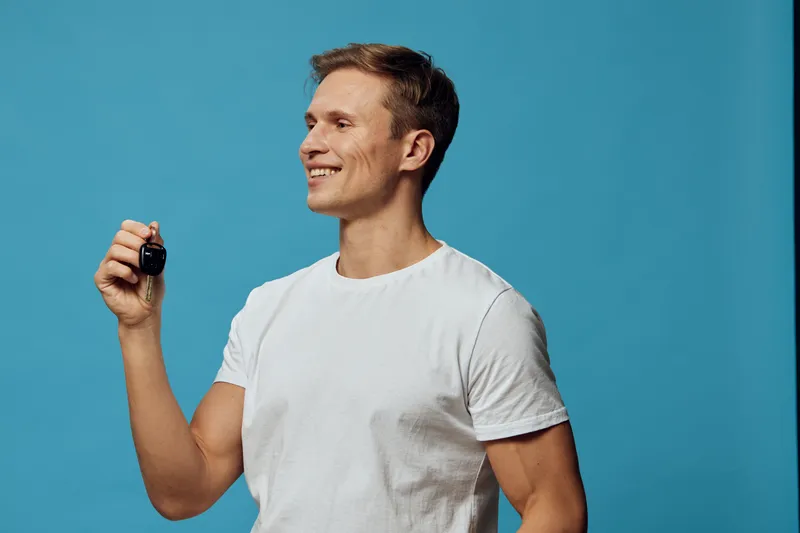 Smiling man holding car key over blue background, casual clothing, cheerful expression, isolated in studio shot, lifestyle concept, human emotions, success, confidence, modern fashion, bright colors.